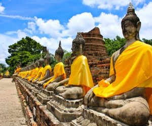 A row of stone Buddha statues, draped in vibrant yellow robes, set against a bright blue sky with scattered clouds.