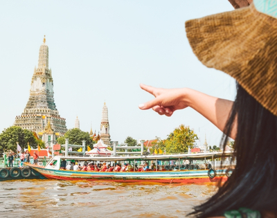 A woman in a straw hat points towards Wat Arun temple while a colorful boat passes by on the Chao Phraya River in Bangkok.