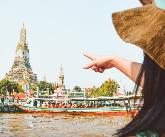 A woman in a straw hat points towards Wat Arun temple while a colorful boat passes by on the Chao Phraya River in Bangkok.