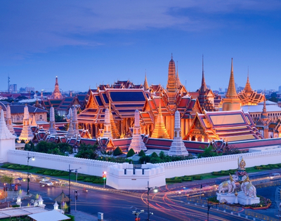 A panoramic view of the Grand Palace in Bangkok, illuminated at twilight with intricate architecture and surrounding gardens.