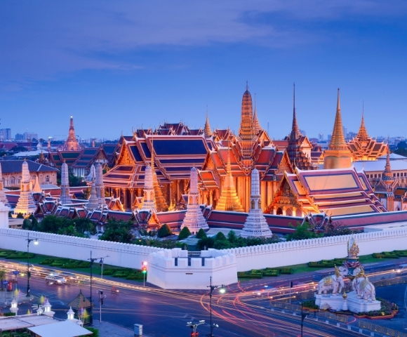 A panoramic view of the Grand Palace in Bangkok, illuminated at twilight with intricate architecture and surrounding gardens.