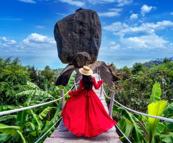 A woman in a flowing red dress and straw hat stands on a bridge, gazing at a giant balanced rock against a vibrant blue sky.