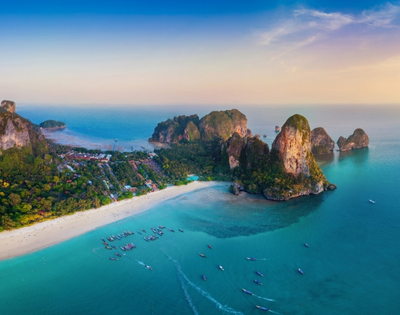 Aerial view of a tropical beach with turquoise water, limestone cliffs, and fishing boats dotting the serene coastline at sunset.