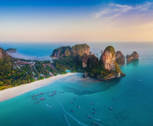 Aerial view of a tropical beach with turquoise water, limestone cliffs, and fishing boats dotting the serene coastline at sunset.
