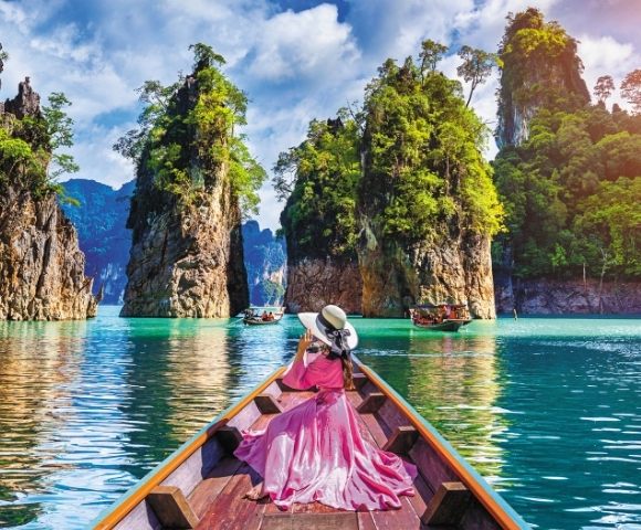 A woman in a pink dress and wide-brimmed hat relaxes on a boat, surrounded by lush islands and turquoise waters.