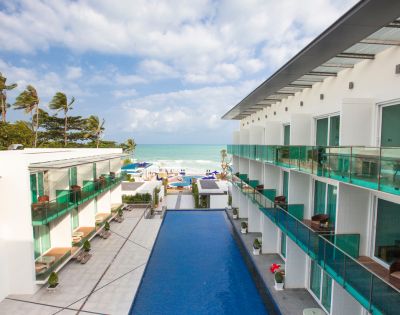 A modern beachfront hotel with turquoise pool, palm trees, and ocean views under a partly cloudy sky.