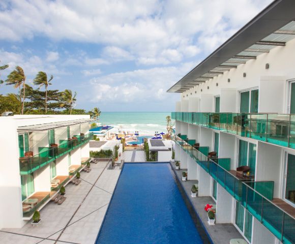 A modern beachfront hotel with turquoise pool, palm trees, and ocean views under a partly cloudy sky.