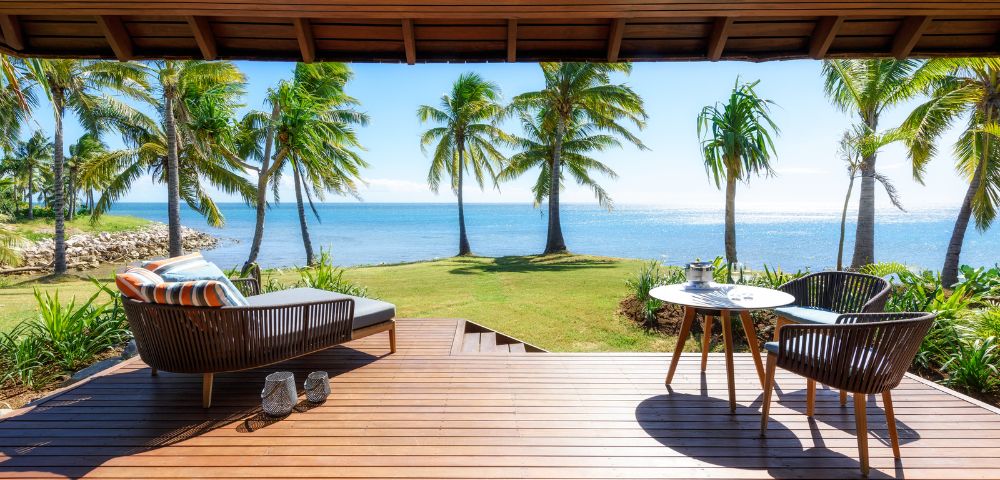 A sunny beachfront view from a wooden deck with palm trees, featuring a lounging chair and a dining table with chairs.