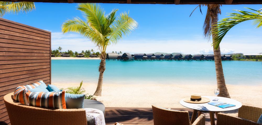 A serene beachfront view from a cozy patio featuring lounge chairs, a table with a hat, and palm trees against a turquoise lagoon.