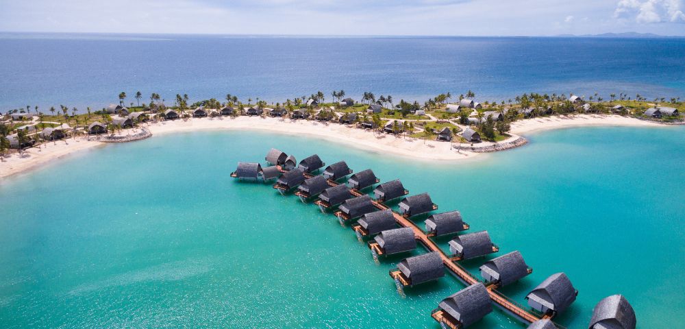 Aerial view of overwater bungalows connected by pathways, surrounded by turquoise water and lush green coastline under a blue sky.
