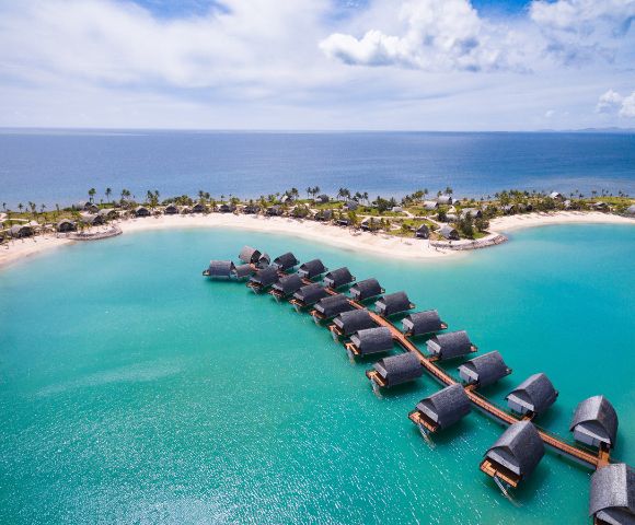 Aerial view of overwater bungalows connected by pathways, surrounded by turquoise water and lush green coastline under a blue sky.