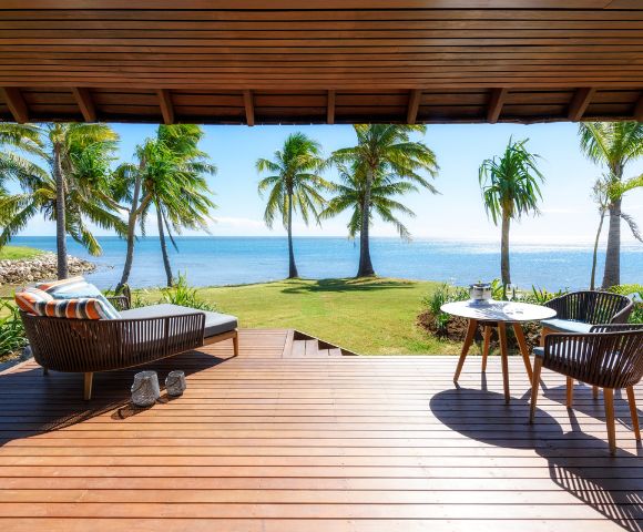 A sunny beachfront view from a wooden deck with palm trees, featuring a lounging chair and a dining table with chairs.