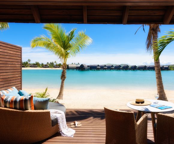 A serene beachfront view from a cozy patio featuring lounge chairs, a table with a hat, and palm trees against a turquoise lagoon.