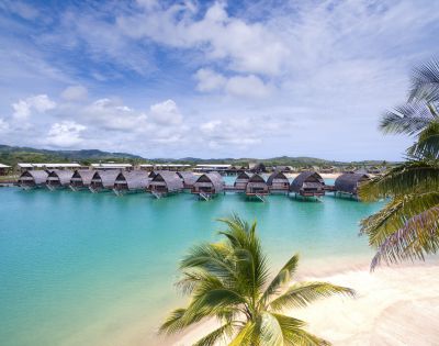 Tropical lagoon with overwater bungalows, clear turquoise water, and sandy beach framed by palm trees under a bright blue sky.
