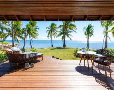 A sunny beachfront view from a wooden deck with palm trees, featuring a lounging chair and a dining table with chairs.