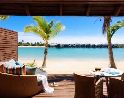 A serene beachfront view from a cozy patio featuring lounge chairs, a table with a hat, and palm trees against a turquoise lagoon.