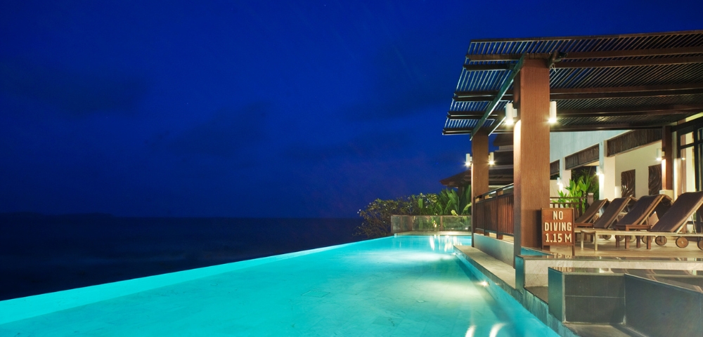 A serene infinity pool at night, illuminated by soft lights, overlooks a calm ocean under a deep blue sky.