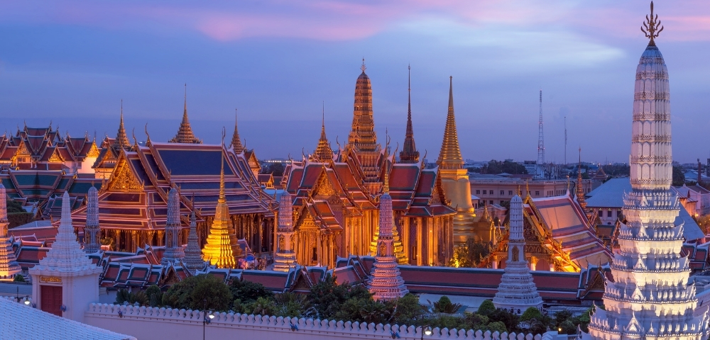 Twilight view of Bangkok's Grand Palace, showcasing golden spires and intricate architecture under a colorful sky.