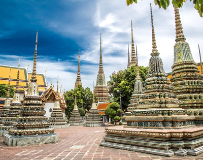 A stunning view of Wat Pho's chedis, adorned with intricate tiles, under a blue sky with fluffy clouds. Lush greenery enhances the serene atmosphere.
