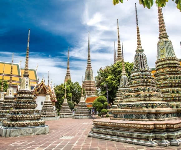 A stunning view of Wat Pho's chedis, adorned with intricate tiles, under a blue sky with fluffy clouds. Lush greenery enhances the serene atmosphere.
