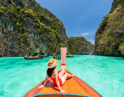 A woman in a red swimsuit lounges on a traditional boat, surrounded by turquoise waters and lush green cliffs. Sunny, tropical paradise.