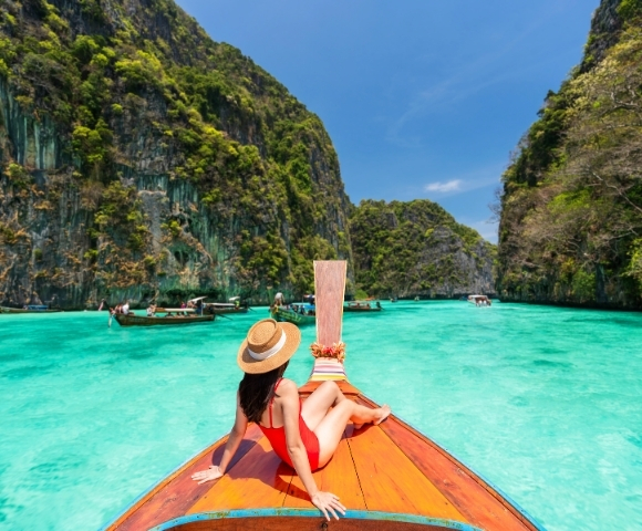 A woman in a red swimsuit lounges on a traditional boat, surrounded by turquoise waters and lush green cliffs. Sunny, tropical paradise.