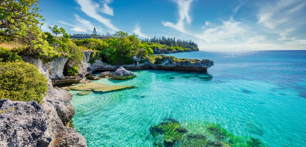 A serene coastal landscape featuring clear turquoise waters, rocky shores, lush greenery, and a bright blue sky with wispy clouds.