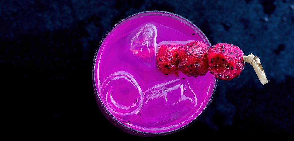A vibrant pink drink with ice, garnished with skewered red fruit, sits on a dark surface.