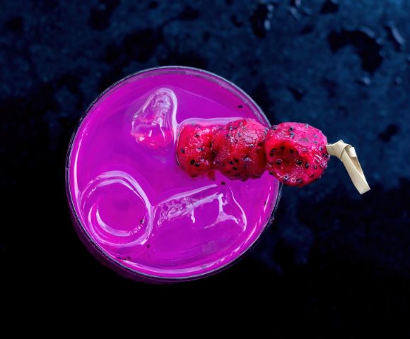 A vibrant pink drink with ice, garnished with skewered red fruit, sits on a dark surface.