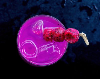 A vibrant pink drink with ice, garnished with skewered red fruit, sits on a dark surface.