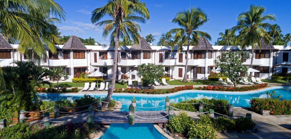 Tropical resort with swimming pool, lush gardens, and palm trees, featuring white buildings with thatched roofs under a clear blue sky.