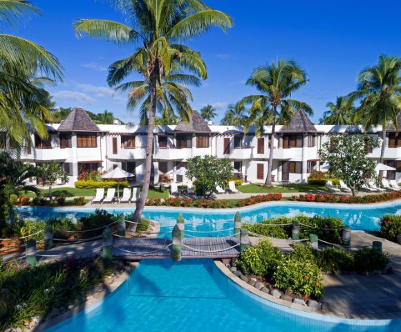 Tropical resort with swimming pool, lush gardens, and palm trees, featuring white buildings with thatched roofs under a clear blue sky.