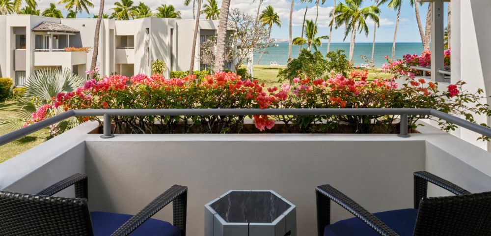 Balcony view with tropical palm trees and colorful flowers, overlooking a beach and clear blue ocean under a partly cloudy sky.