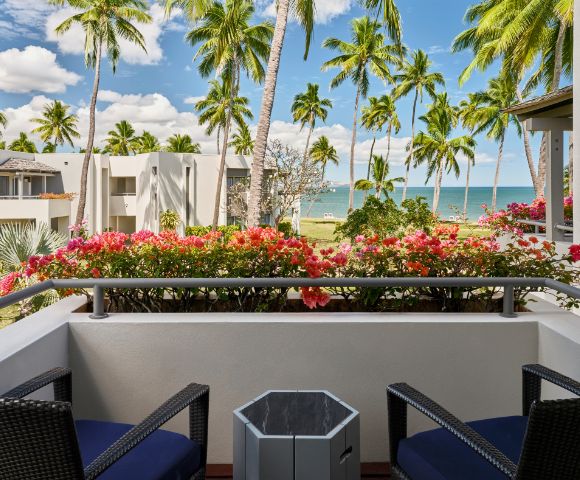 Balcony view with tropical palm trees and colorful flowers, overlooking a beach and clear blue ocean under a partly cloudy sky.
