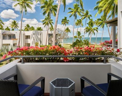 Balcony view with tropical palm trees and colorful flowers, overlooking a beach and clear blue ocean under a partly cloudy sky.