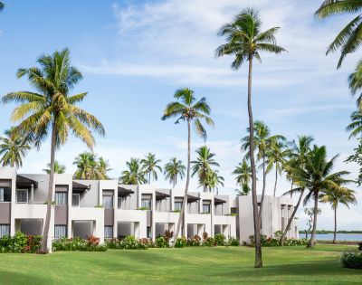 Tropical resort with modern white buildings surrounded by lush green lawns and palm trees against a clear blue sky.