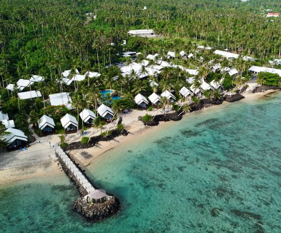 Aerial view of a beachfront resort with white-roofed bungalows amidst lush greenery, ocean, and a pier extending into clear waters.