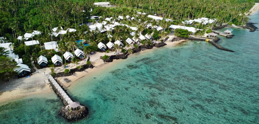 Aerial view of a beachfront resort with white-roofed bungalows amidst lush greenery, ocean, and a pier extending into clear waters.