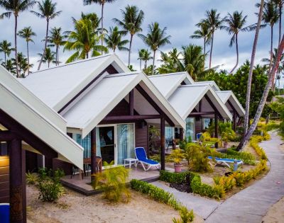 A row of tropical bungalows with sloped roofs, surrounded by lush greenery and palm trees, along a sandy walkway.
