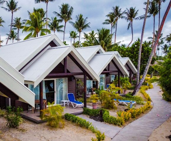 A row of tropical bungalows with sloped roofs, surrounded by lush greenery and palm trees, along a sandy walkway.