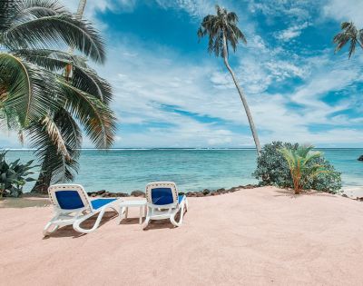 Two lounge chairs on a sandy beach, surrounded by palm trees, overlooking a tranquil turquoise sea and a blue sky with scattered clouds.