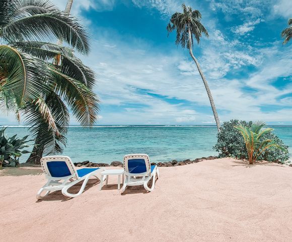 Two lounge chairs on a sandy beach, surrounded by palm trees, overlooking a tranquil turquoise sea and a blue sky with scattered clouds.