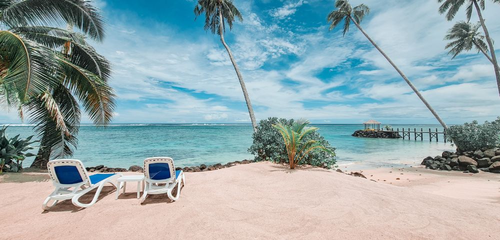 Two lounge chairs on a sandy beach, surrounded by palm trees, overlooking a tranquil turquoise sea and a blue sky with scattered clouds.