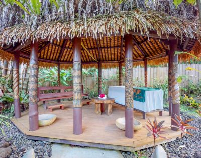 Tropical gazebo with a thatched roof, wooden benches, and a small table, surrounded by lush greenery and rocks.