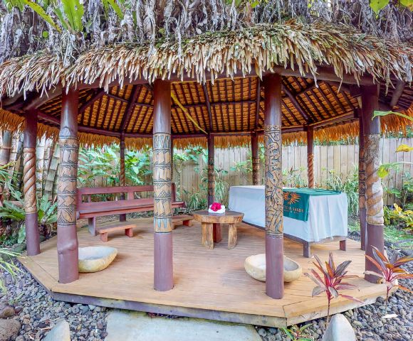 Tropical gazebo with a thatched roof, wooden benches, and a small table, surrounded by lush greenery and rocks.