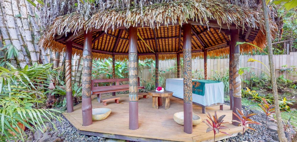 Tropical gazebo with a thatched roof, wooden benches, and a small table, surrounded by lush greenery and rocks.