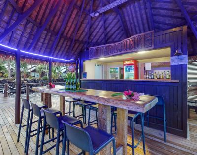 Tropical bar scene with a thatched roof, wooden counter, and high stools, featuring drinks and vibrant decor in a relaxing setting.