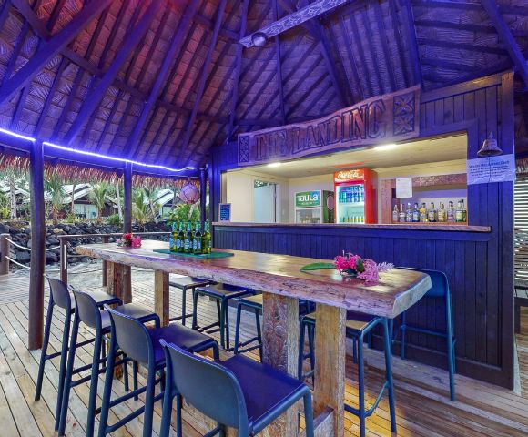 Tropical bar scene with a thatched roof, wooden counter, and high stools, featuring drinks and vibrant decor in a relaxing setting.