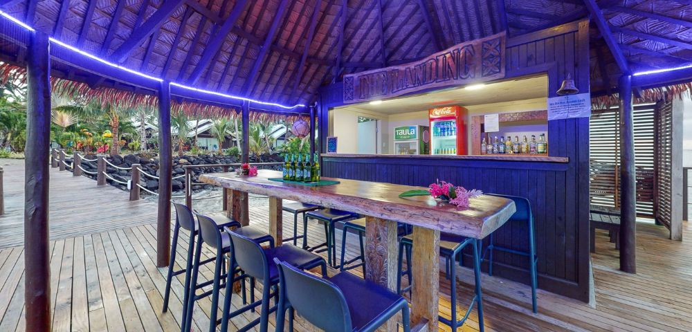 Tropical bar scene with a thatched roof, wooden counter, and high stools, featuring drinks and vibrant decor in a relaxing setting.