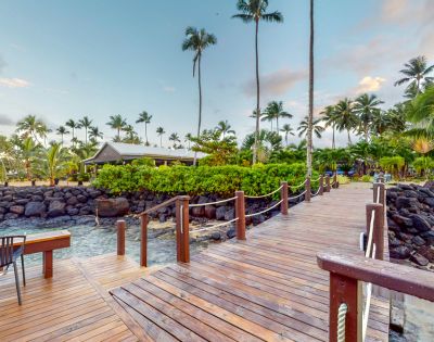 A wooden walkway leads through lush greenery and palm trees towards a serene waterside setting. A tranquil atmosphere invites relaxation.
